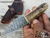 Stag Handle Damascus Knife with a hand-forged raindrop pattern blade, featuring a genuine sambar stag antler handle and a cowhide leather sheath, displayed on a rustic wooden background.