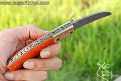 Handmade Damascus Steel Folding Knife with a hawkbill blade, engraved brass bolsters, and an orange camel bone handle. The knife comes with a genuine cowhide leather sheath.