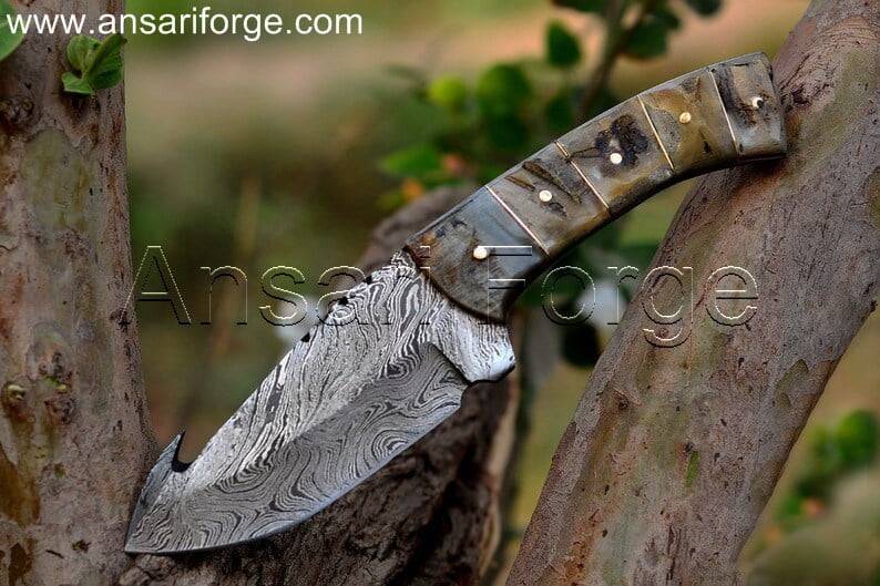 Hand-forged Damascus steel hunting knife with gut hook and ram horn handle for efficient field dressing.