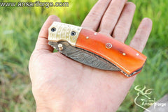 Handmade Damascus Steel Folding Knife with a hawkbill blade, engraved brass bolsters, and an orange camel bone handle. The knife comes with a genuine cowhide leather sheath.
