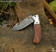 Handmade Damascus pocket knife resting on a wooden surface in a forest setting, illustrating its suitability for outdoor adventures and nature exploration.